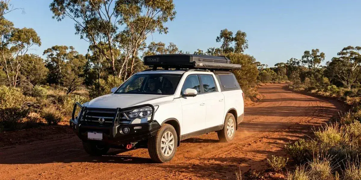 A 4WD dual-cab camper with rooftop tent travelling along a red unsealed outback road surrounded by Australian bush landscape in Western Australia