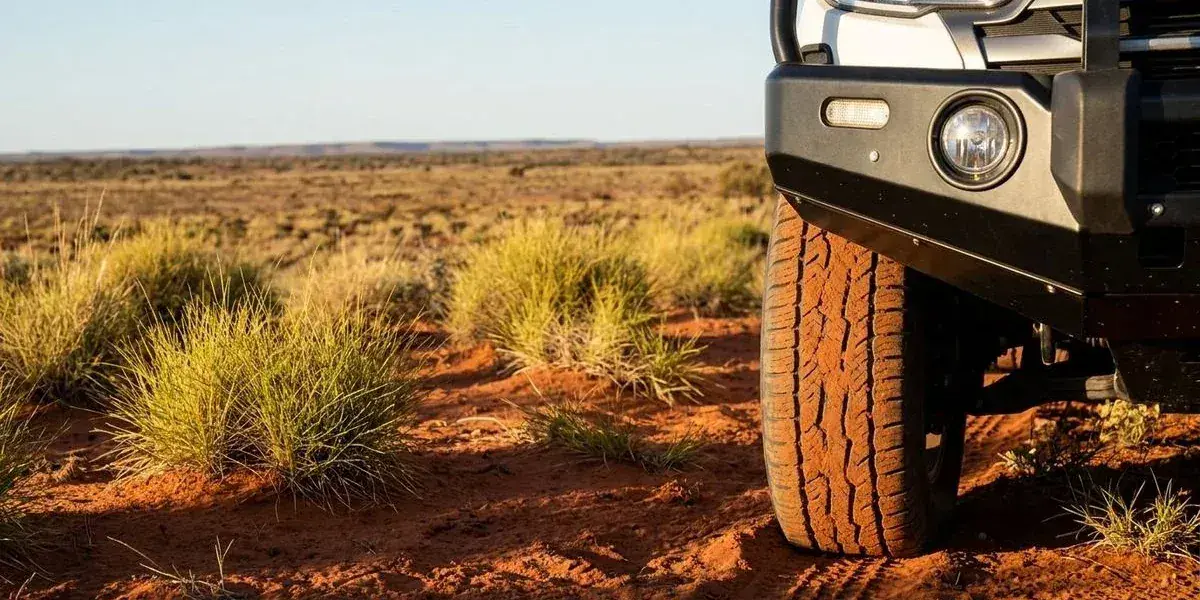 A close-up of 4WD tyres on red sandy terrain in the Western Australian outback with spinifex grass and a distant horizon showing the remote landscape