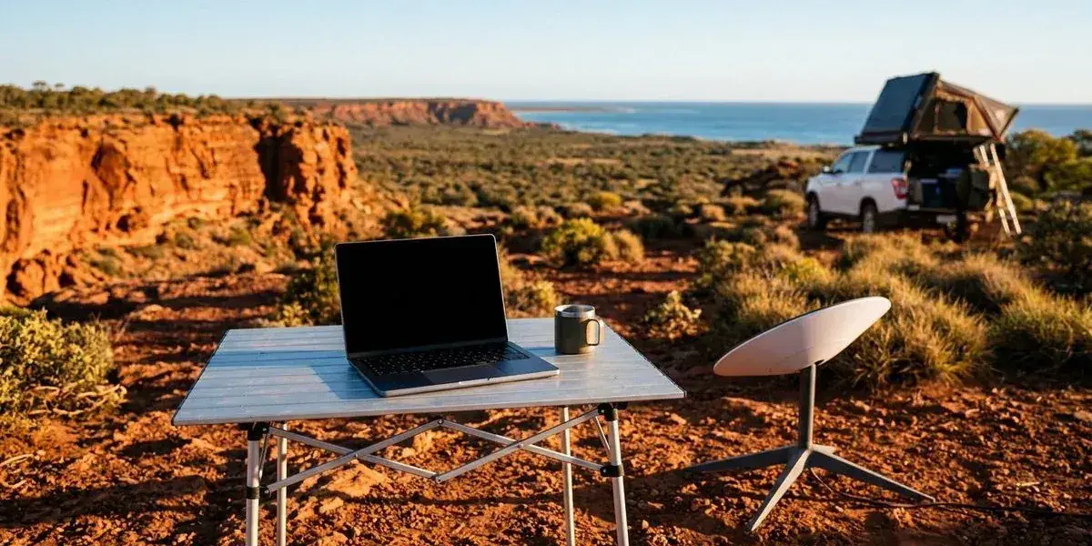 Laptop open on a camp table with Starlink dish visible nearby at a remote Western Australian campsite overlooking red cliffs and bush landscape