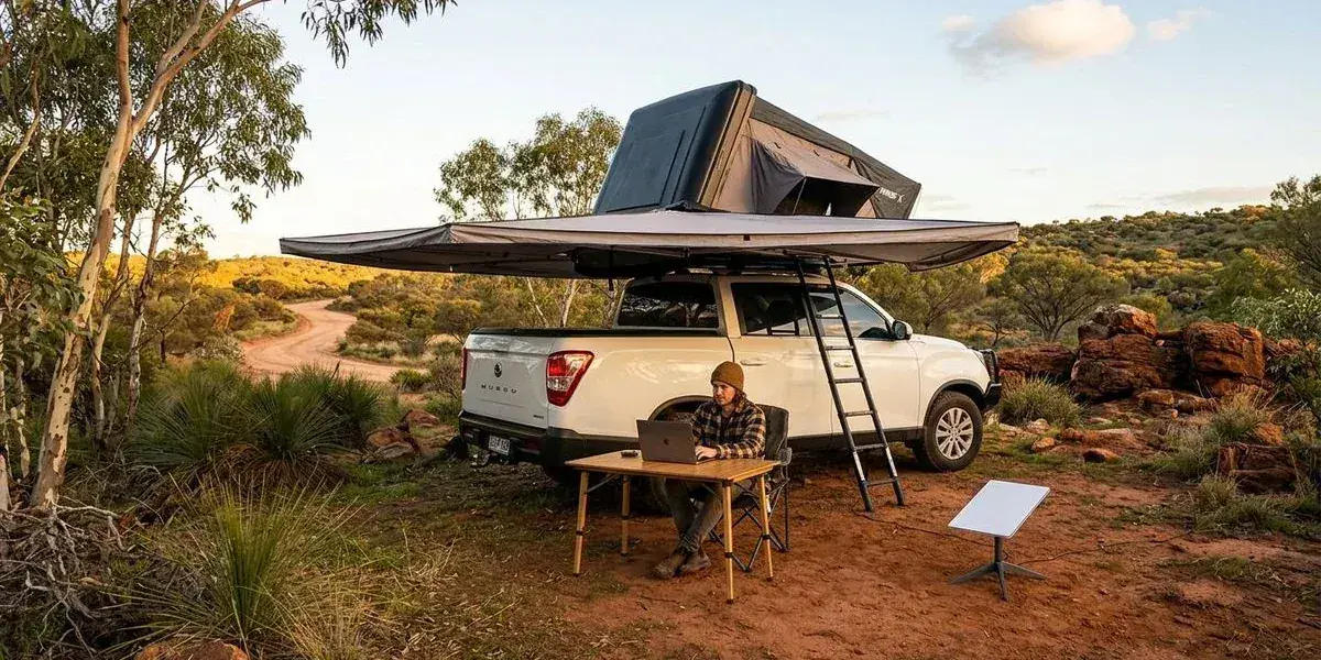 A person sitting under a 270 degree awning attached to a 4WD camper working on a laptop with a Starlink dish nearby at a remote Western Australian bush campsite