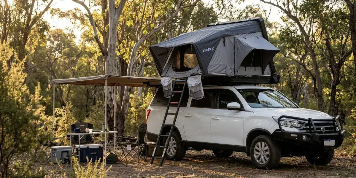 A spacious campsite setup with a dual-cab 4WD camper showing deployed awning camp table and chairs with bushland in the background